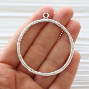 May include: A silver-colored metal circle pendant with a hammered texture and a small loop at the top. The pendant is held in a hand, showcasing its design and potential for jewelry making or crafting projects.
