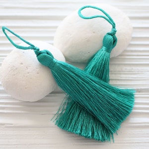 May include: Two teal blue tassels with long, flowing strands. The tassels are on a white background.