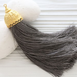 May include: A gray tassel with a gold metal top. The tassel is made of soft, silky threads and has a decorative, ornate top.