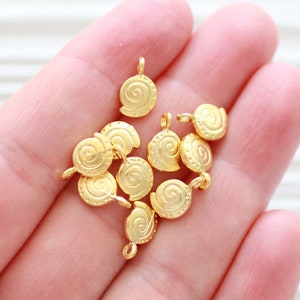 May include: A close-up of a hand holding a pile of gold-colored metal charms. Each charm is a small, round disc with a raised spiral design.
