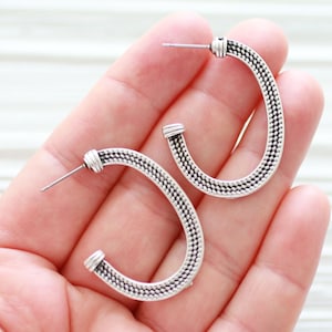 May include: A pair of silver hoop earrings with a textured oval design. The earrings are shown on a hand.