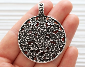 May include: Silver pendant with a circular design featuring a delicate floral pattern. The pendant is made of metal and has a loop at the top for attaching to a chain.