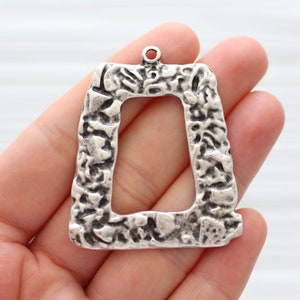 May include: Silver-toned metal pendant with a rectangular frame and a textured surface. The frame has a rough, uneven texture.