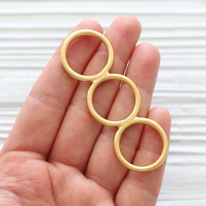 May include: A gold-toned metal ring with three interlocking circles. The ring is held in a hand, with the white background of a wooden surface visible.