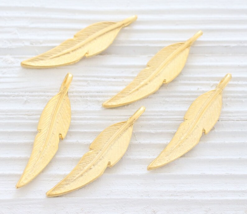 May include: Five gold-colored metal feather charms. The feathers are all the same size and shape, with a slightly textured surface.