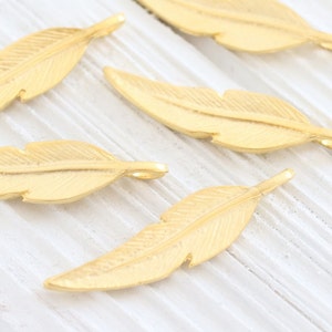 May include: Five gold-colored metal feather charms with a textured surface. The charms have a hole at the top for attaching to jewelry or other crafts.