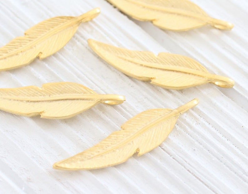 May include: Five gold-colored metal feather charms with a textured surface. The charms have a hole at the top for attaching to jewelry or other crafts.