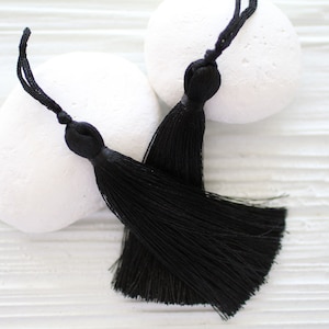 May include: Two black tassel ornaments with long, thin strands. The tassels are on a white background.