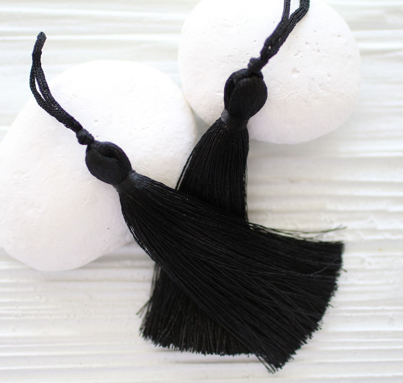 May include: Two black tassel ornaments with long, thin strands. The tassels are on a white background.