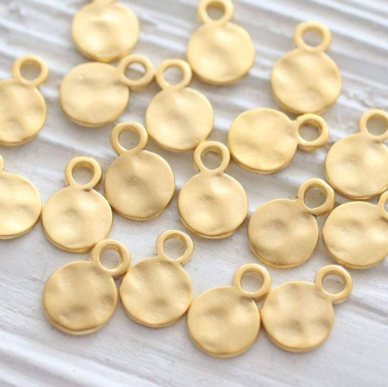 May include: A collection of 15 gold-toned metal circle charms with a textured surface and a double loop at the top. The charms are scattered on a white surface.
