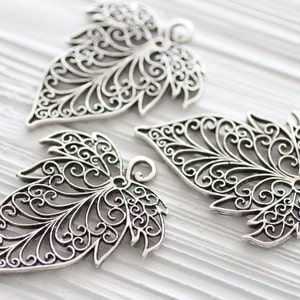 Op de afbeelding: Drie zilveren filigrein bladeren-charms met ingewikkelde ontwerpen. De charms zijn delicaat en sierlijk, met een gedetailleerd patroon van spiralen en bochten.