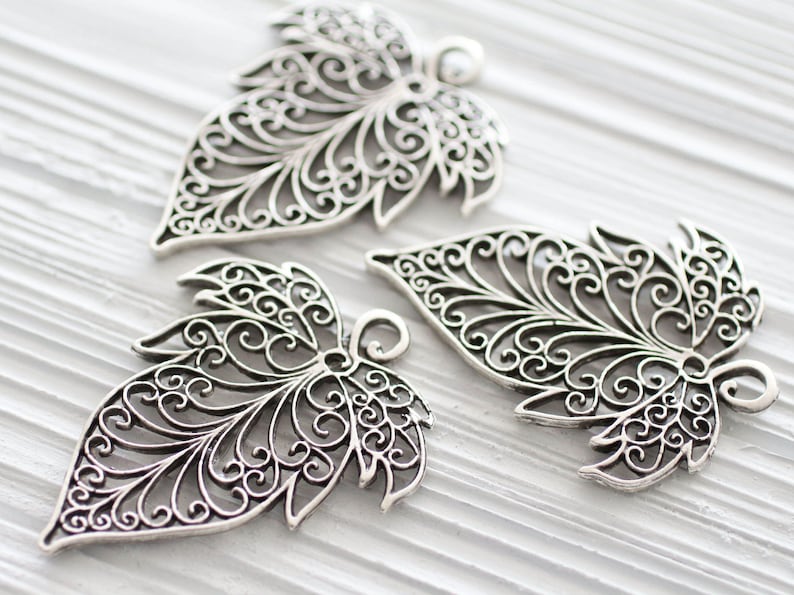 Op de afbeelding: Drie zilveren filigrein bladeren-charms met ingewikkelde ontwerpen. De charms zijn delicaat en sierlijk, met een gedetailleerd patroon van spiralen en bochten.
