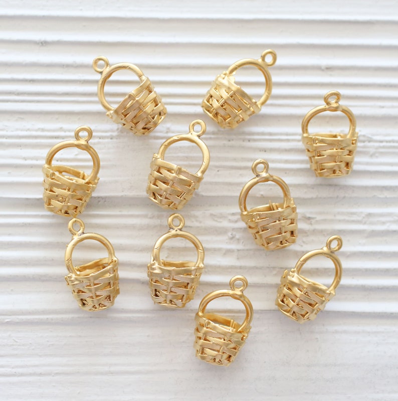 May include: Ten gold-colored metal basket charms with a woven design. The charms have a loop at the top for attaching to jewelry or other crafts.