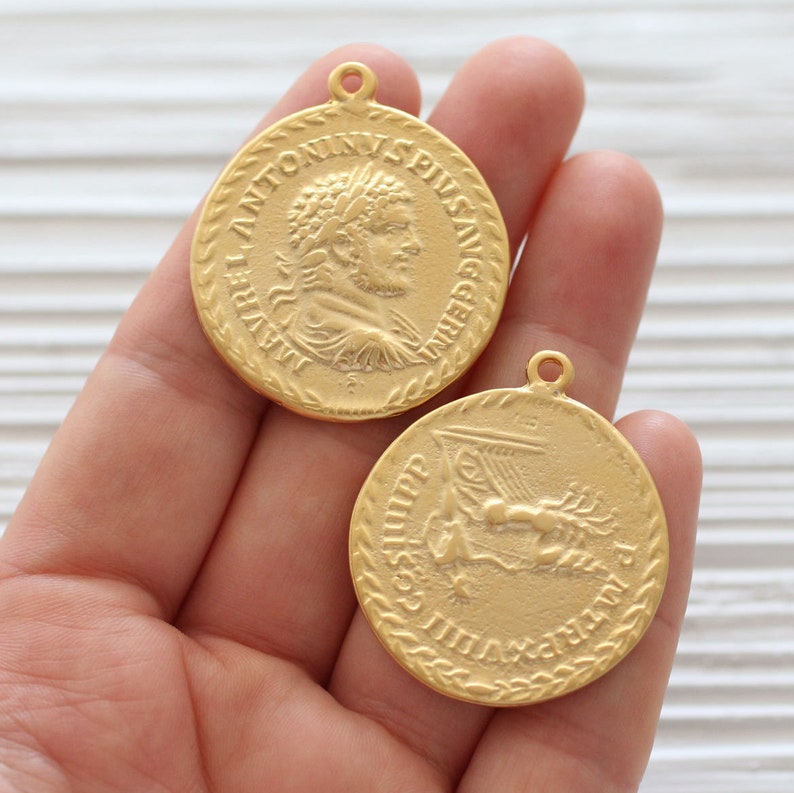 May include: Two gold-toned metal charms, one with a profile of a Roman emperor with the inscription "LAVRENTIVS ANTONINVS PIVS AVG", and the other with a ship and the inscription "CESAR IMP CAESAR AVG F", both with a rope-like border.