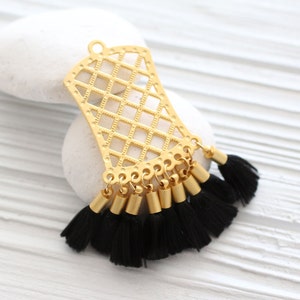 May include: A gold-colored filigree pendant with a diamond pattern and a series of black tassels. The pendant has a loop at the top for hanging. The tassels are attached to gold-colored cylindrical beads.
