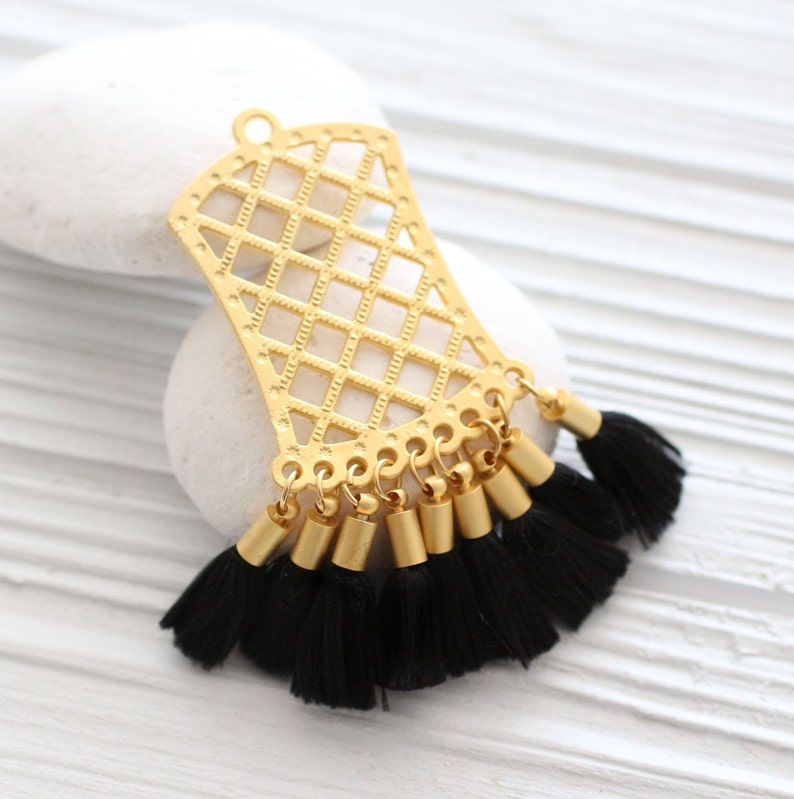 May include: A gold-colored filigree pendant with a diamond pattern and a series of black tassels. The pendant has a loop at the top for hanging. The tassels are attached to gold-colored cylindrical beads.
