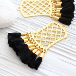 May include: Two gold-colored filigree pendants with black tassels. The pendants have a geometric lattice design and are connected to multiple black tassels with gold-colored caps. The background is a white, textured surface.