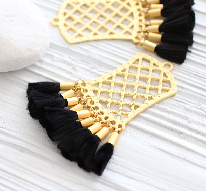 May include: Two gold-colored filigree pendants with black tassels. The pendants have a geometric lattice design and are connected to multiple black tassels with gold-colored caps. The background is a white, textured surface.