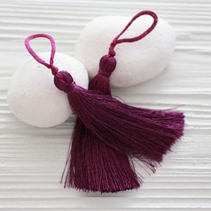 May include: Two plum-coloured tassels with looped cords, resting on white, smooth stones. The tassels have a bulbous knot at the top and long, flowing strands. The background is a white, textured surface.