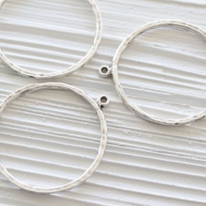 May include: Three silver-toned metal hoop charms with a hammered texture. Each charm has a small loop at the top for attaching to other jewelry components. The hoops are circular and have a rustic, handmade appearance, suitable for jewelry making.