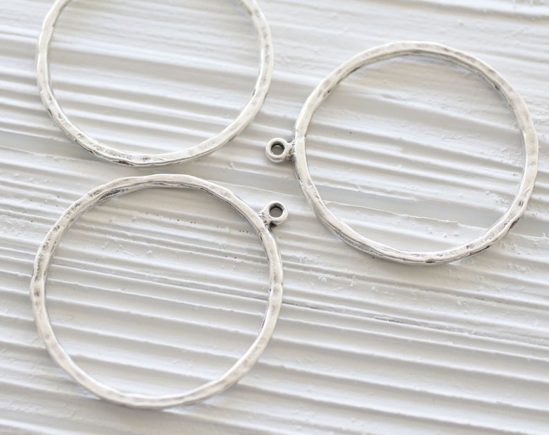 May include: Three silver-toned metal hoop charms with a hammered texture. Each charm has a small loop at the top for attaching to other jewelry components. The hoops are circular and have a rustic, handmade appearance, suitable for jewelry making.