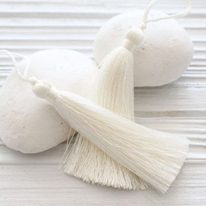 May include: Two white tassel trim embellishments on a white background. The tassels are made of soft, fluffy fibers and are perfect for adding a touch of elegance to any project.