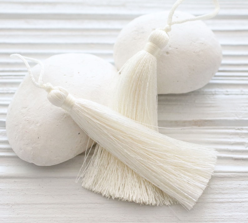 May include: Two white tassel trim embellishments on a white background. The tassels are made of soft, fluffy fibers and are perfect for adding a touch of elegance to any project.