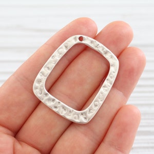 May include: A silver-colored, hammered metal pendant with a square shape and rounded corners. The pendant has a small hole at the top for stringing. The surface has a textured, hammered appearance.