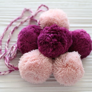 Puede incluir: Un grupo de pompones esponjosos de color rosa y morado atados juntos con una cuerda blanca y rosa.