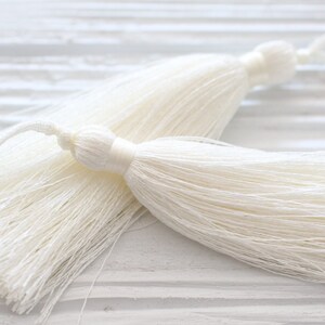 May include: Two white tassel trims with a long, thin, and fluffy texture. The tassels are made of a soft, silky material and have a delicate, elegant appearance.