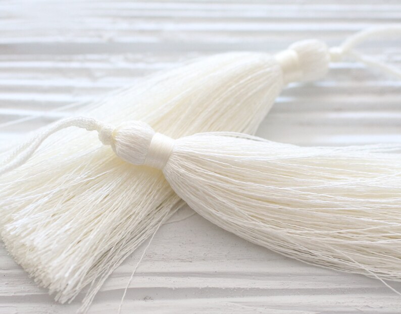 May include: Two white tassel trims with a long, thin, and fluffy texture. The tassels are made of a soft, silky material and have a delicate, elegant appearance.