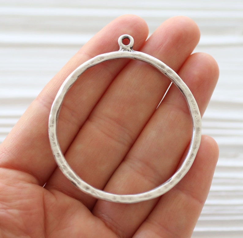 May include: A silver-colored metal circle pendant with a hammered texture and a small loop at the top. The pendant is held in a hand, showcasing its design and potential for jewelry making or crafting projects.