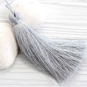 May include: A light gray tassel with a looped top and long, flowing strands. The tassel is lying on a white, textured surface with two white, rounded stones. The tassel is a decorative element.