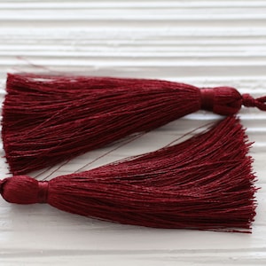 May include: Two burgundy tassel trims with a knotted cord. The tassels are made of soft, fine threads and have a long, flowing fringe.