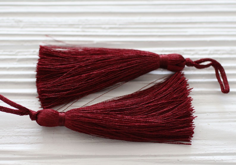 May include: Two burgundy tassel trims with a knotted cord. The tassels are made of soft, fine threads and have a long, flowing fringe.