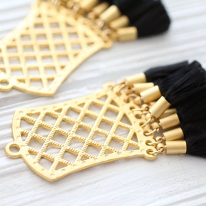 May include: Gold-colored filigree pendants with attached black tassels. The pendants have a geometric lattice design and small loops for attaching to jewelry. The tassels are made of black thread and have gold-colored metal caps.