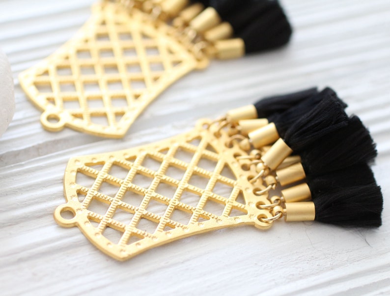 May include: Gold-colored filigree pendants with attached black tassels. The pendants have a geometric lattice design and small loops for attaching to jewelry. The tassels are made of black thread and have gold-colored metal caps.