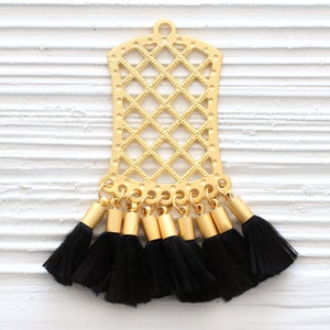 May include: Gold-colored metal pendant with a lattice design and a series of black tassels. The pendant has a rectangular shape with a rounded top and a small loop for hanging. The tassels are attached to the bottom.