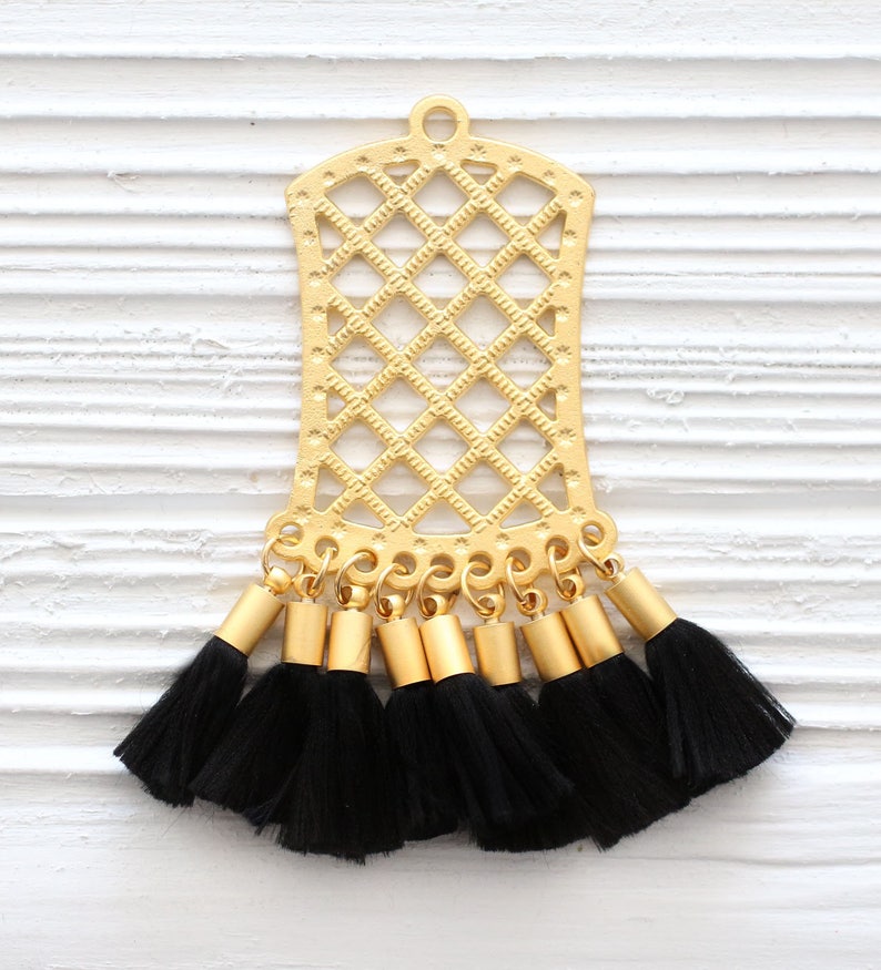 May include: Gold-colored metal pendant with a lattice design and a series of black tassels. The pendant has a rectangular shape with a rounded top and a small loop for hanging. The tassels are attached to the bottom.
