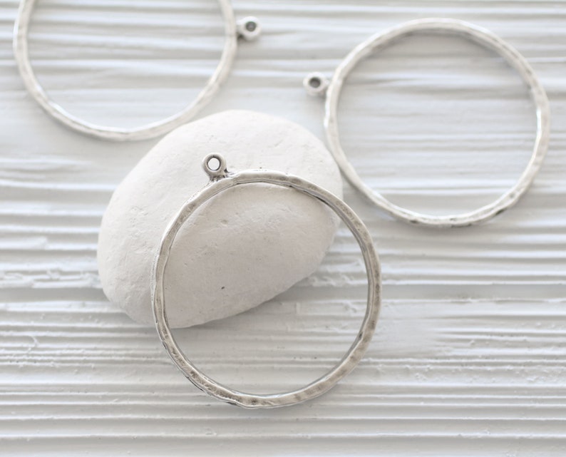 May include: Silver-toned metal circle pendants with a hammered texture. Each pendant has a small loop at the top for attaching to a chain or other jewelry component. The pendants are displayed on a white surface.