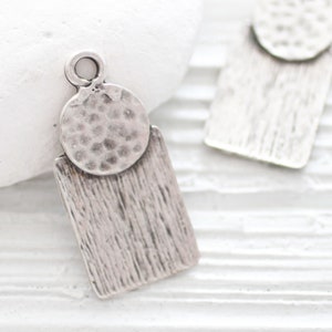 May include: Silver charm with a textured round disc and a rectangular pendant. The charm has a hammered finish and a loop for attaching to a necklace or bracelet.