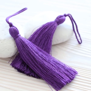 May include: Two purple tassel ornaments with long strands of fringe. The tassels are tied together and are lying on a white surface.