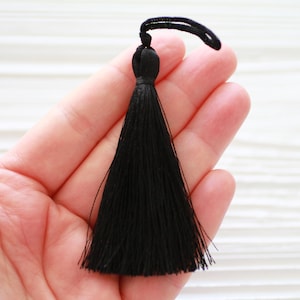 May include: A black tassel with a long, flowing fringe. The tassel is held in a hand, and the background is a white wooden surface.