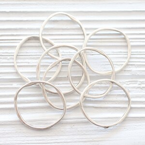 May include: A set of seven silver rings, each with a slightly different shape and size, arranged in a pile on a white wooden surface.