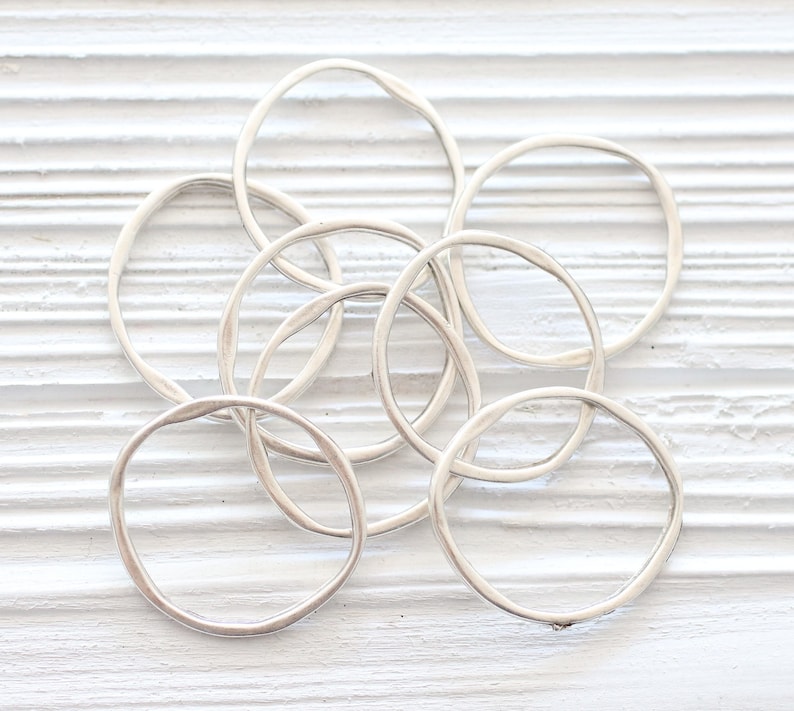 May include: A set of seven silver rings, each with a slightly different shape and size, arranged in a pile on a white wooden surface.
