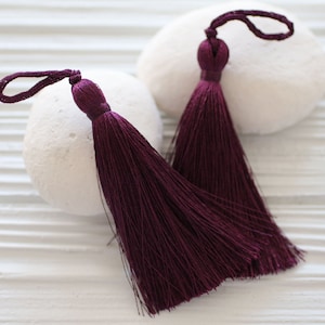 May include: Two dark purple silk tassels with a loop at the top. The tassels are on a white background.