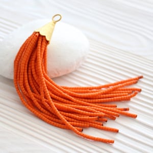 May include: Orange tassel with a gold metal top. The tassel is made of many small beads and is hanging from a white surface.