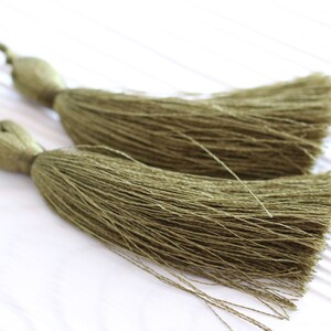 May include: Two olive green tassel ornaments with long, thin strands of fabric. The tassels are tied together at the top.