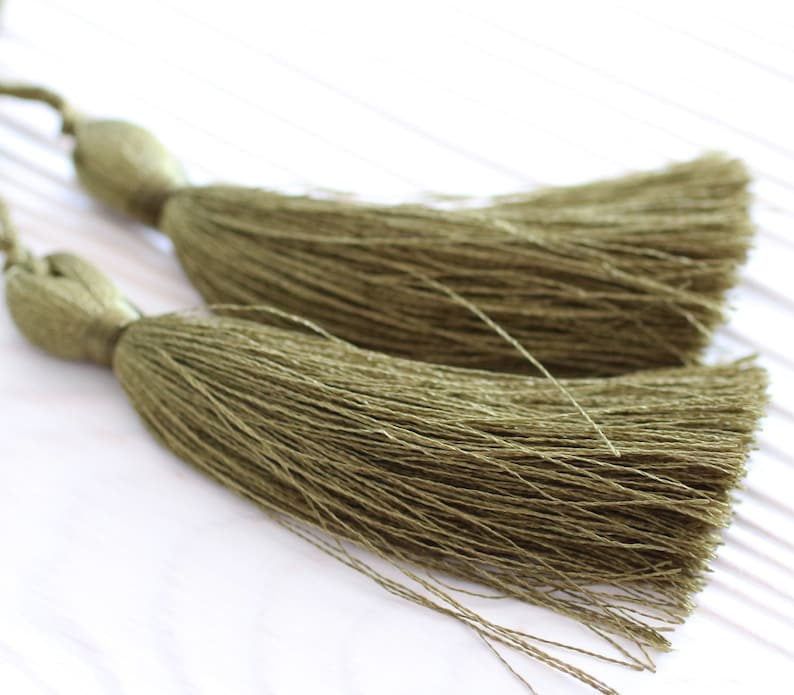 May include: Two olive green tassel ornaments with long, thin strands of fabric. The tassels are tied together at the top.