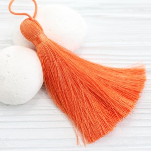 May include: Close-up of an orange tassel with a loop, resting on two white, smooth stones. The tassel is made of fine, silky threads, and the background is a white, textured surface. The tassel is a decorative element.